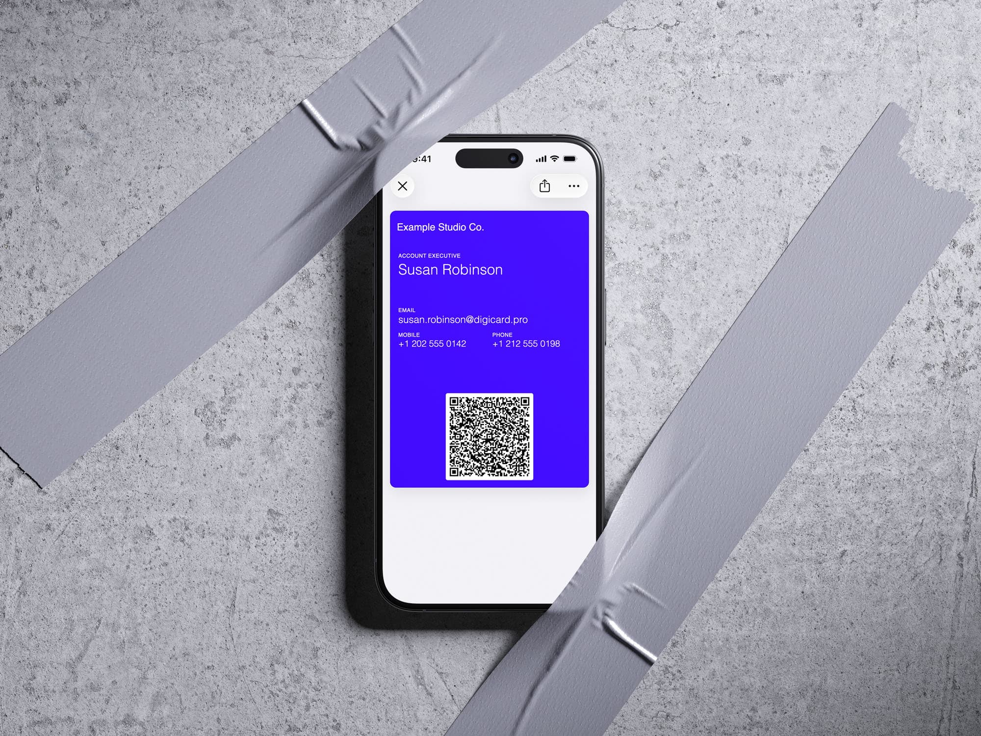 iPhone taped to a concrete wall showing an indigo Apple Wallet business card with contact details and a QR code