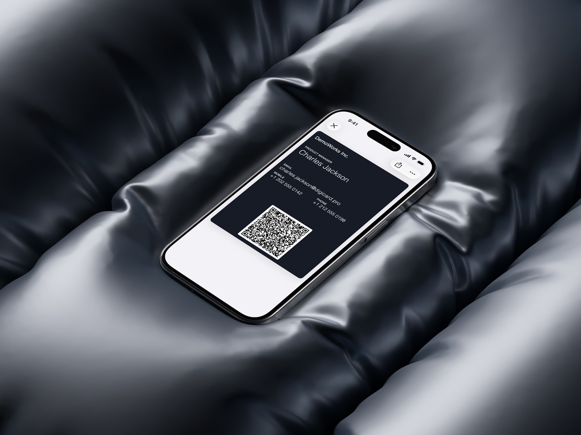 iPhone resting on glossy black quilted fabric, screen showing a dark-navy Apple Wallet digital business card for Charles Jackson with email, phone numbers, and a scannable QR code