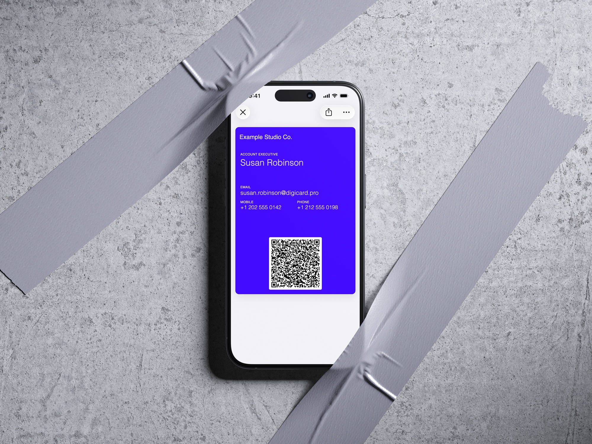 iPhone taped to a concrete wall showing an indigo Apple Wallet business card with contact details and a QR code