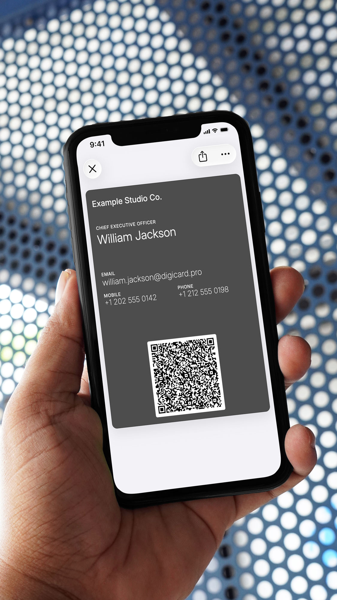 A hand holds an iPhone showing a graphite-gray digital business card in Apple Wallet for William Jackson, Chief Executive Officer at Example Studio Co., with email, mobile number, and a QR code, against a blue perforated-metal background